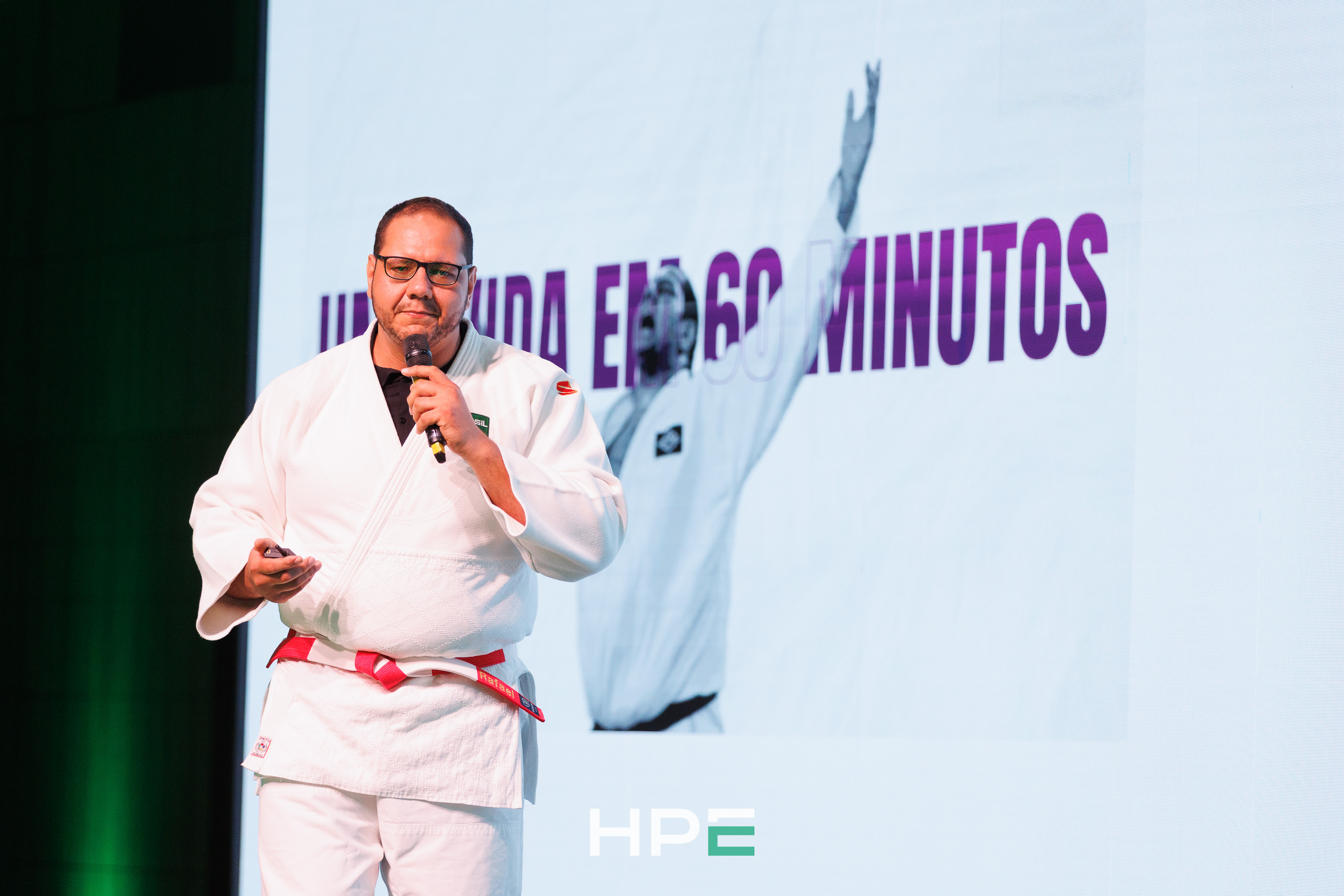 Rafael Silva de kimono e faixa vermelha no palco, microfone na mão, diante do telão da palestra Uma Vida em 60 Minutos