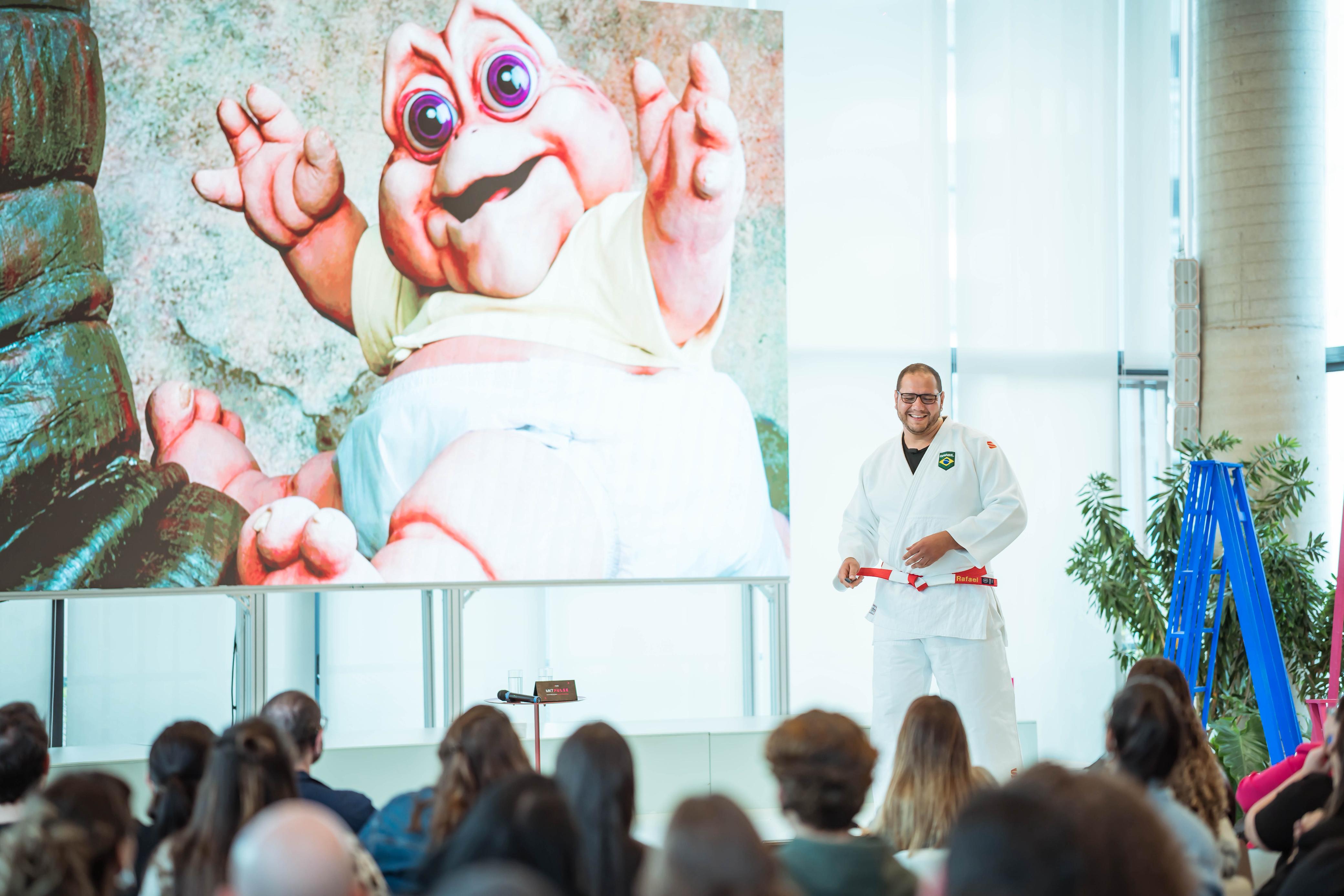 Rafael Silva de kimono no palco, diante do telão com a imagem do bebê da Família Dinossauro, para uma plateia sentada